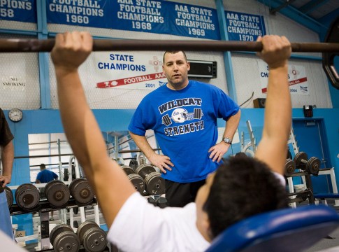 A high-school football player lifts weights while his coach watches.